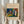 May include: Person in cream clothing holding framed abstract art print featuring vibrant blues, greens, yellows, reds and white brushstrokes in Kandinsky's expressionist style. Beige textured mat with gallery text, light wood frame, dried pampas grass visible to left, neutral interior setting with white curtains and soft natural lighting.