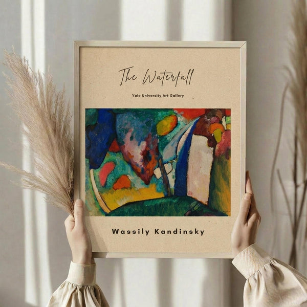 May include: Person in cream clothing holding framed abstract art print featuring vibrant blues, greens, yellows, reds and white brushstrokes in Kandinsky's expressionist style. Beige textured mat with gallery text, light wood frame, dried pampas grass visible to left, neutral interior setting with white curtains and soft natural lighting.