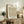 May include: Framed botanical poster featuring William Morris daisy pattern with green leaves and orange-red flowers, displayed on rustic wooden sideboard alongside fern plant, books, ceramic vase and beige upholstered chair in warm-toned living room setting