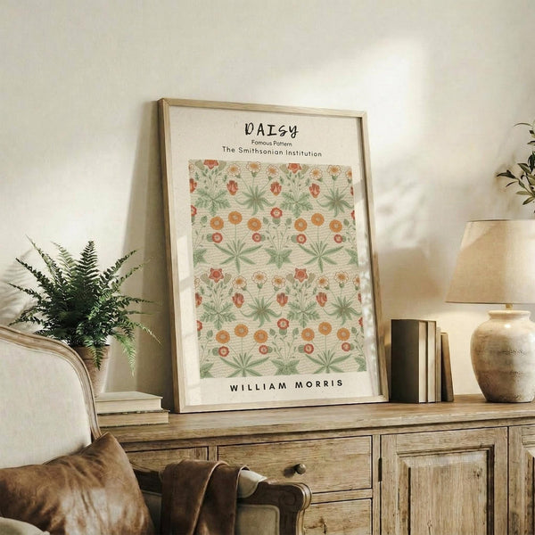 May include: Framed botanical poster featuring William Morris daisy pattern with green leaves and orange-red flowers, displayed on rustic wooden sideboard alongside fern plant, books, ceramic vase and beige upholstered chair in warm-toned living room setting