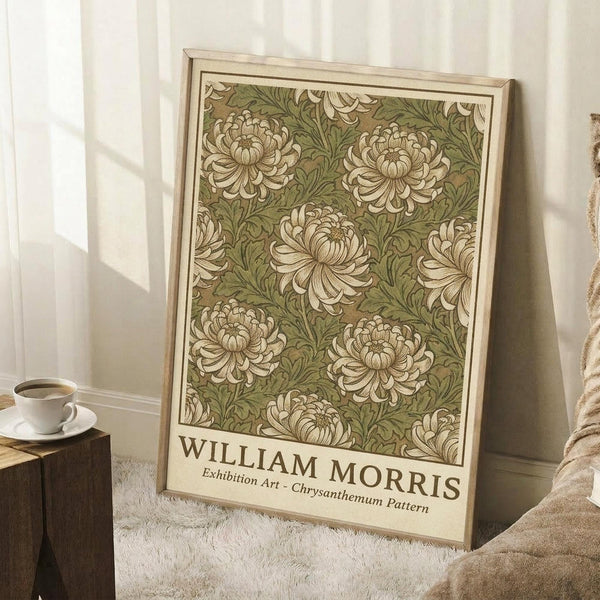 May include: Framed botanical art print featuring cream chrysanthemum flowers on sage green background, displayed in light wood frame leaning against white wall corner. Scene shows cozy setting with coffee cup on wooden surface and soft beige throw blanket nearby, natural window lighting creating gentle shadows on white walls.