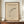 May include: Abstract minimalist poster with geometric black curves and circles on beige background, displayed in brown wooden frame on walnut credenza. White ceramic vase and stacked books flank the artwork. Clean modern interior with white wainscoting walls and natural lighting creates sophisticated gallery-like setting.