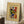 May include: Framed abstract art poster leaning against white wall corner on white textured rug. Colorful Kandinsky-style painting with swirling brushstrokes in green, red, blue, yellow and black on cream background with exhibition text. Light wood frame, cozy room setting with coffee cup on wooden block and beige blanket visible.