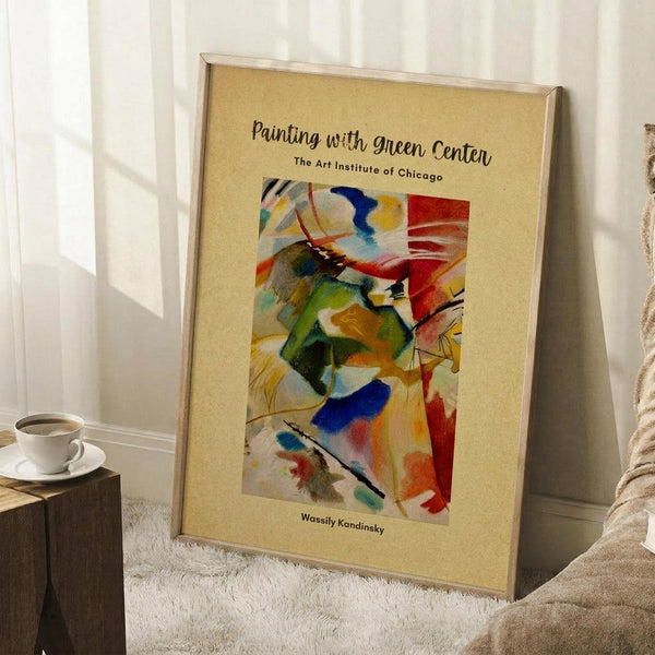 May include: Framed abstract art poster leaning against white wall corner on white textured rug. Colorful Kandinsky-style painting with swirling brushstrokes in green, red, blue, yellow and black on cream background with exhibition text. Light wood frame, cozy room setting with coffee cup on wooden block and beige blanket visible.