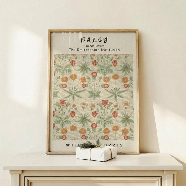 May include: Framed botanical art print featuring William Morris daisy pattern with green foliage and orange-red flowers on cream background, displayed on light wood console table beside white wrapped gift with evergreen sprig, against neutral wall in minimalist home setting
