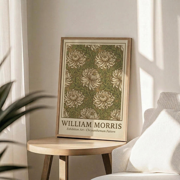 May include: Framed botanical art print featuring cream chrysanthemum flowers on olive green background, displayed on round wooden side table next to beige upholstered chair. Natural lighting creates soft shadows across neutral-toned room with white curtains and plant leaves visible in corner.