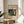 May include: Framed art poster featuring an abstract expressionist painting with vibrant blues, greens, reds, and yellows in geometric and organic shapes. The print has cream-colored margins with "The Waterfall," "Yale University Art Gallery," and "Wassily Kandinsky" text. Displayed in thin wooden frame on round wooden side table next to beige upholstered chair with plant leaves visible, soft natural lighting creating shadows on white wall.