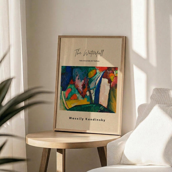 May include: Framed art poster featuring an abstract expressionist painting with vibrant blues, greens, reds, and yellows in geometric and organic shapes. The print has cream-colored margins with "The Waterfall," "Yale University Art Gallery," and "Wassily Kandinsky" text. Displayed in thin wooden frame on round wooden side table next to beige upholstered chair with plant leaves visible, soft natural lighting creating shadows on white wall.