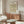 May include: Framed botanical poster with green daisy pattern hanging above wooden sideboard in bright living room. Person's hand visible on credenza, white curved chair and glass coffee table in foreground. Warm natural lighting creates shadows on cream walls.
