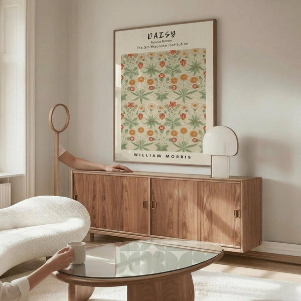 May include: Framed botanical poster with green daisy pattern hanging above wooden sideboard in bright living room. Person's hand visible on credenza, white curved chair and glass coffee table in foreground. Warm natural lighting creates shadows on cream walls.