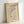 May include: Framed minimalist art print featuring abstract black curved lines and geometric shapes including circles and a dot on cream-colored paper, displayed in light wood frame leaning against white wall on light surface