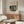 May include: Abstract colorful poster print featuring vibrant blues, greens, reds, and yellows in Kandinsky's expressionist style, displayed in thin wooden frame above walnut credenza. Modern living room setting with cream curved chair, glass oval coffee table, and white table lamp. Warm natural lighting creates shadows across minimalist neutral decor.