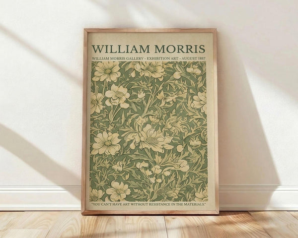 May include: Framed vintage William Morris exhibition poster featuring intricate green and cream floral pattern with flowing botanical designs. The Arts and Crafts style artwork shows detailed flowers, leaves and vines against sage green background. Wooden frame leans against white wall on light wood floor with soft natural lighting creating diagonal shadows across the scene.