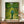 May include: Framed Van Gogh poster print displayed on wooden credenza against white paneled wall. Artwork shows figure in blue shirt and green pants walking down path between fruit trees with yellow oranges. Post-impressionist style with swirling brushstrokes in vibrant greens and yellows. Person's hands visible adjusting frame. White ceramic vase and books nearby for scale.