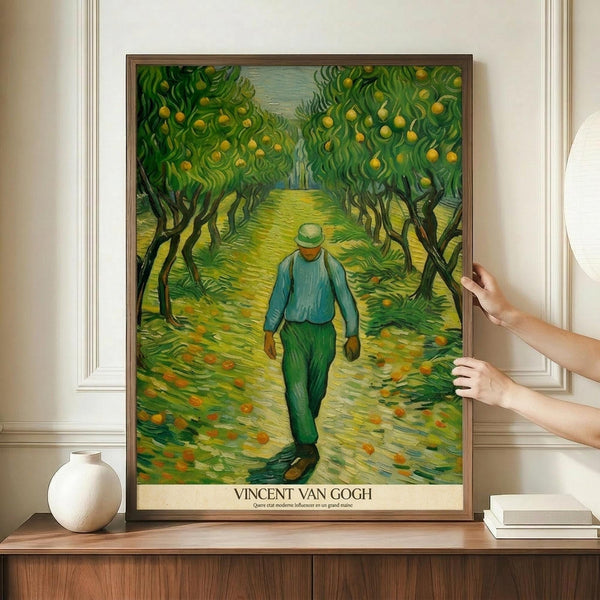 May include: Framed Van Gogh poster print displayed on wooden credenza against white paneled wall. Artwork shows figure in blue shirt and green pants walking down path between fruit trees with yellow oranges. Post-impressionist style with swirling brushstrokes in vibrant greens and yellows. Person's hands visible adjusting frame. White ceramic vase and books nearby for scale.
