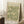 May include: Framed William Morris-style botanical poster with sage green and cream floral pattern featuring detailed flowers and foliage, displayed in light wooden frame leaning against white wainscoted wall beside textured throw blanket and wooden side table with white coffee cup and saucer.