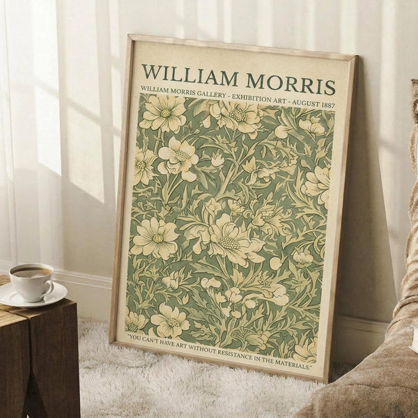 May include: Framed William Morris-style botanical poster with sage green and cream floral pattern featuring detailed flowers and foliage, displayed in light wooden frame leaning against white wainscoted wall beside textured throw blanket and wooden side table with white coffee cup and saucer.