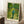 May include: Framed Van Gogh poster print leaning against white wall on textured cream rug. Artwork shows figure in blue shirt and green pants walking down path through orchard with fruit trees and fallen oranges scattered on ground. Swirling brushstroke style in vibrant greens and yellows. Natural wood frame with cream matting. Coffee cup on wooden side table nearby, soft natural lighting creating shadows.