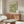 May include: Vintage botanical poster with William Morris floral pattern in sage green and cream tones, displayed above mid-century walnut credenza in bright modern living room. White curved chair and glass oval coffee table visible, with minimalist copper accent lighting and neutral wall colors.
