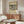 May include: Modern living room interior featuring a framed vintage art poster on beige wall above wooden sideboard. Abstract black line drawing on cream background shows dynamic figures and forms. Room styled with white curved chair, glass oval coffee table, white table lamp, and copper floor lamp on polished floors.