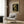 May include: Van Gogh's skeleton with burning cigarette poster displayed on wood paneled wall above beige upholstered armchair with geometric patterned pillow. Modern brass floor lamp and round black side table with open book nearby. Vintage-style museum poster features cream background with bold black text and classic skull painting image.