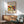 May include: Framed abstract art poster with vibrant red, yellow, blue, and white geometric shapes and figures, displayed above a light wood credenza with white ceramic lamp. Mint green mid-century armchair with wooden frame sits nearby in sunlit room with white walls and warm shadows across wooden floor.