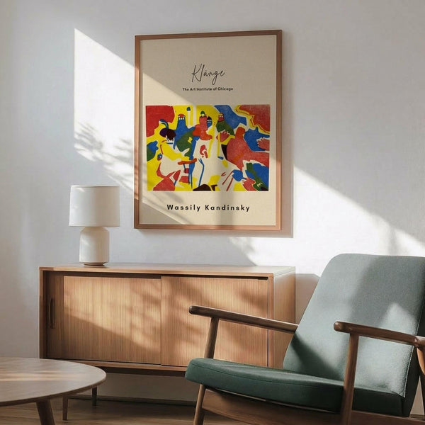 May include: Framed abstract art poster with vibrant red, yellow, blue, and white geometric shapes and figures, displayed above a light wood credenza with white ceramic lamp. Mint green mid-century armchair with wooden frame sits nearby in sunlit room with white walls and warm shadows across wooden floor.