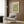 May include: Cream-colored vintage botanical poster with green floral pattern mounted on wood paneled wall above modern beige upholstered chair with geometric pillow. Scene shows contemporary reading nook with brass floor lamp, black side table with open book, and natural lighting from window with white curtains.