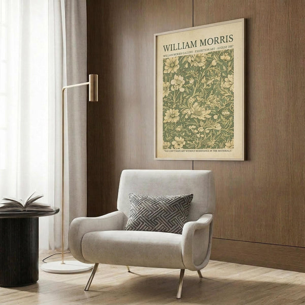 May include: Cream-colored vintage botanical poster with green floral pattern mounted on wood paneled wall above modern beige upholstered chair with geometric pillow. Scene shows contemporary reading nook with brass floor lamp, black side table with open book, and natural lighting from window with white curtains.