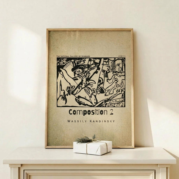 May include: Framed vintage-style poster featuring Wassily Kandinsky's abstract black and white artwork from 1911, displayed on light wood surface with small wrapped gift box. The print shows bold, flowing abstract forms and is titled "Composition 2" with museum attribution text. Natural wood frame against cream wall in minimalist setting.