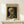May include: Framed Van Gogh museum poster featuring a skeleton smoking a cigarette against dark background, displayed on light wood surface next to small white wrapped gift with green twine. The vintage-style print shows cream and brown tones in a natural wood frame, leaning against white wall.