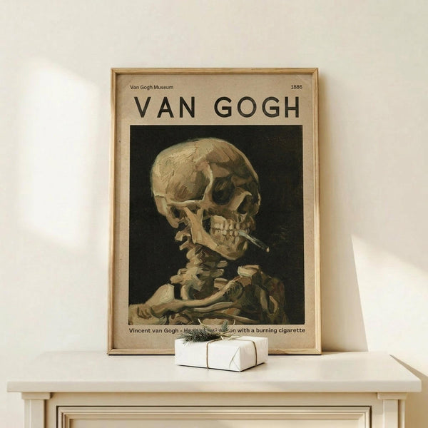 May include: Framed Van Gogh museum poster featuring a skeleton smoking a cigarette against dark background, displayed on light wood surface next to small white wrapped gift with green twine. The vintage-style print shows cream and brown tones in a natural wood frame, leaning against white wall.