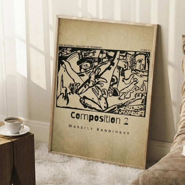 May include: A framed beige poster featuring Wassily Kandinsky's abstract black line drawing from 1911, displayed against white paneled walls. The artwork shows flowing organic forms and bold strokes in a monochromatic style. A coffee cup sits on a wooden block nearby, with a cream-colored throw blanket visible on a white textured rug.