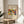 May include: Framed art print with abstract colorful composition featuring bold red, yellow, blue, and green shapes with stylized figures. Print shows cursive "Klänge" text at top, "The Art Institute of Chicago" below, and "Wassily Kandinsky" at bottom. Displayed in thin wooden frame on round wooden side table next to beige upholstered chair, with plant leaves visible and natural sunlight casting shadows on cream wall.