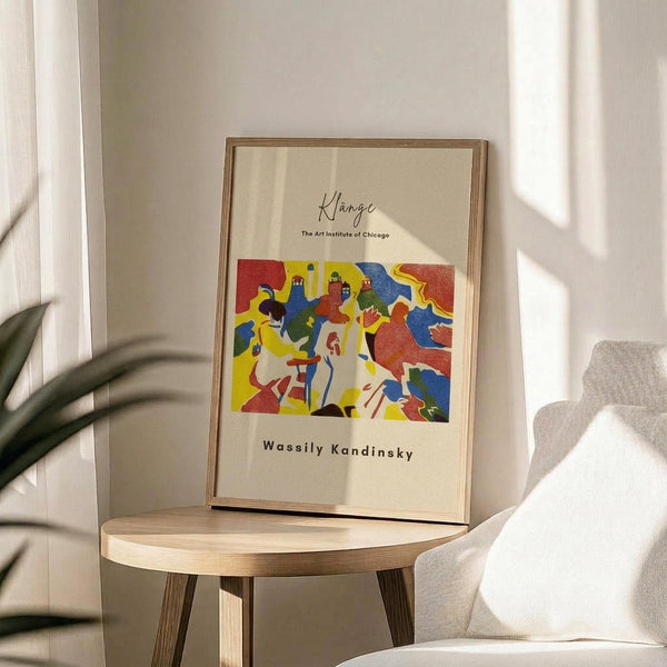 May include: Framed art print with abstract colorful composition featuring bold red, yellow, blue, and green shapes with stylized figures. Print shows cursive "Klänge" text at top, "The Art Institute of Chicago" below, and "Wassily Kandinsky" at bottom. Displayed in thin wooden frame on round wooden side table next to beige upholstered chair, with plant leaves visible and natural sunlight casting shadows on cream wall.