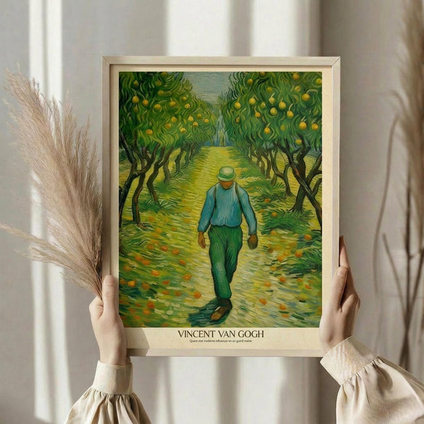 May include: Hands holding a framed Van Gogh reproduction print showing a figure in blue shirt and green pants walking through an orchard with yellow fruit trees. The artwork features Van Gogh's signature swirling brushstroke style in vibrant greens and yellows. Light wooden frame against white curtained background with dried pampas grass visible on the left side.