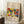 May include: A framed art poster leaning against a white wall in a cozy room setting. The print features vibrant abstract figures in bold red, yellow, blue, and green colors with text reading "Klänge" and "The Art Institute of Chicago" above and "Wassily Kandinsky" below. The poster has a light wooden frame and sits on a white textured rug next to a wooden side table with a white coffee cup and saucer. Soft natural light creates gentle shadows on the wall.