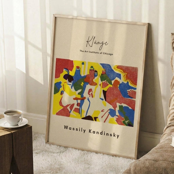 May include: A framed art poster leaning against a white wall in a cozy room setting. The print features vibrant abstract figures in bold red, yellow, blue, and green colors with text reading "Klänge" and "The Art Institute of Chicago" above and "Wassily Kandinsky" below. The poster has a light wooden frame and sits on a white textured rug next to a wooden side table with a white coffee cup and saucer. Soft natural light creates gentle shadows on the wall.