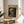 May include: Framed museum poster featuring Van Gogh's painting of a skeleton smoking a cigarette, displayed on a round wooden side table next to a beige upholstered chair. Natural sunlight streams across white walls, creating soft shadows. A plant's green leaves are visible in the lower left corner. The poster has a thin wooden frame and shows a skull with burning cigarette against a dark background.