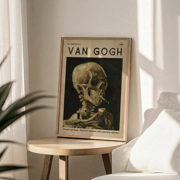 May include: Framed museum poster featuring Van Gogh's painting of a skeleton smoking a cigarette, displayed on a round wooden side table next to a beige upholstered chair. Natural sunlight streams across white walls, creating soft shadows. A plant's green leaves are visible in the lower left corner. The poster has a thin wooden frame and shows a skull with burning cigarette against a dark background.