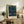 May include: Framed William Morris exhibition poster featuring ornate pattern with birds and foliage in deep blues and greens, displayed on rustic wooden sideboard alongside fern plant, ceramic vase, and books in neutral-toned living room setting with upholstered chair visible.