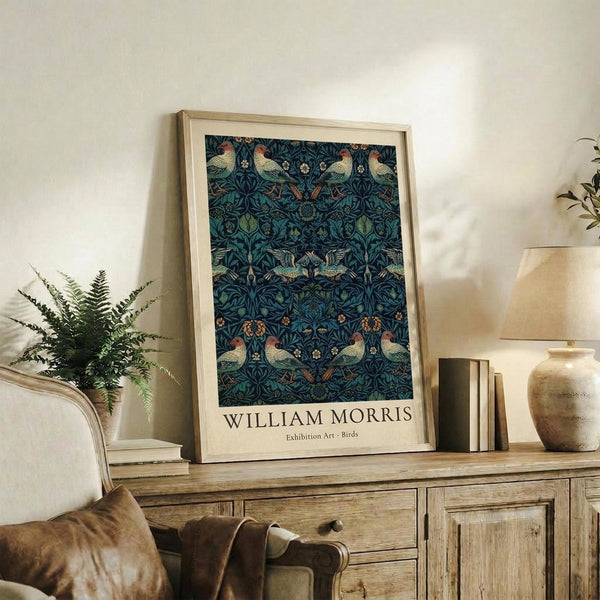 May include: Framed William Morris exhibition poster featuring ornate pattern with birds and foliage in deep blues and greens, displayed on rustic wooden sideboard alongside fern plant, ceramic vase, and books in neutral-toned living room setting with upholstered chair visible.
