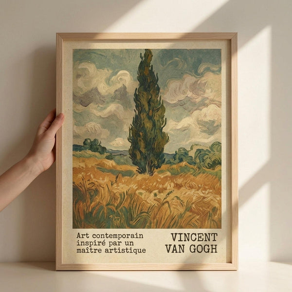 May include: Framed art print in light wood frame held against white wall with natural lighting. Artwork shows Van Gogh-style landscape with tall dark cypress tree in center, golden wheat field below, swirling clouds above in blues and whites. Text at bottom reads "Art contemporain inspiré par un maître artistique" and "VINCENT VAN GOGH". Hand visible holding left side of frame for scale reference.