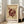 May include: Framed abstract art print in light wood frame leaning against white wall next to beige upholstered sofa. Poster features geometric design with burgundy circular and linear elements on cream background, with museum and artist text. Natural lighting from nearby windows creates soft shadows on hardwood floor.