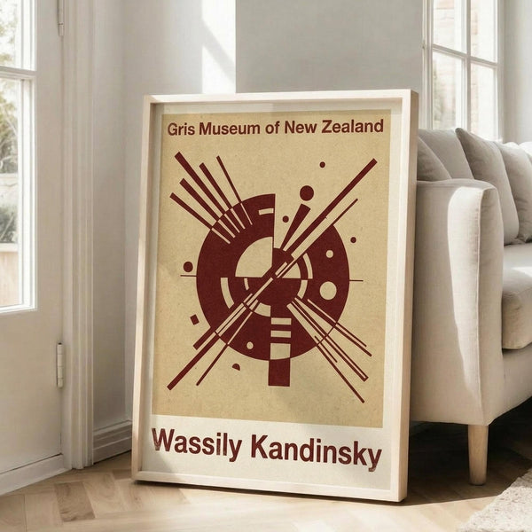 May include: Framed abstract art print in light wood frame leaning against white wall next to beige upholstered sofa. Poster features geometric design with burgundy circular and linear elements on cream background, with museum and artist text. Natural lighting from nearby windows creates soft shadows on hardwood floor.