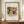May include: Framed poster print of Wassily Kandinsky's abstract expressionist painting featuring flowing organic shapes in earth tones of ochre, burgundy, green and black against cream background. The vintage-style print shows "Improvisation 27 (1912)" text below the artwork. Wooden frame sits on mid-century walnut credenza beside white ceramic vase with bare branches and stack of books against cream walls with white crown molding.