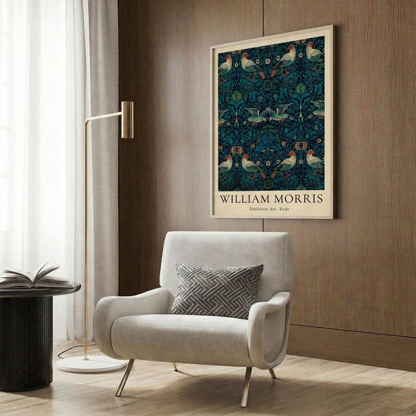 May include: Framed William Morris exhibition poster featuring intricate blue-green pattern with birds and botanical motifs, displayed above a cream upholstered armchair with geometric pillow. Modern room setting with brass floor lamp, dark round side table with open book, wood paneled walls, and natural lighting from window with white curtains.
