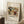 May include: A framed art poster leaning against a white wall corner, featuring Kandinsky's abstract painting with flowing organic shapes in earth tones - greens, browns, yellows, and reds. The artwork shows characteristic curved lines and biomorphic forms on a light background. Below the image is text reading "Improvisation 27" and "(1912)". A white coffee cup sits on a wooden block nearby, with soft natural lighting creating shadows on the textured white rug.