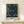 May include: Framed poster featuring William Morris-style pattern with birds and botanical motifs in deep blues and greens, displayed on light wood furniture beside small wrapped gift with twine and greenery sprig against cream wall