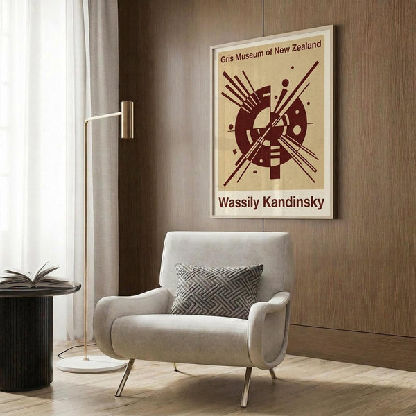 May include: Framed abstract art poster featuring burgundy geometric shapes, circles, and radiating lines on cream background, displayed above a beige upholstered armchair with geometric patterned pillow. Modern living room setting with wood paneled wall, brass floor lamp, and dark round side table with open book.